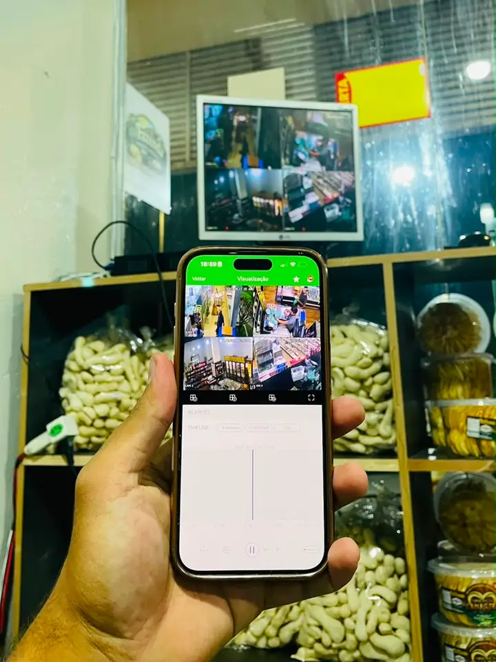 Celular exibindo ao vivo as câmeras de um comércio, com o monitor ao fundo